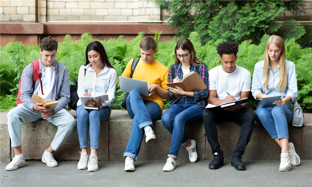 Sechs junge Menschen sitzen draußen auf einer Steinbank mit Schreibheften und Laptops, vermutlich Studenten.