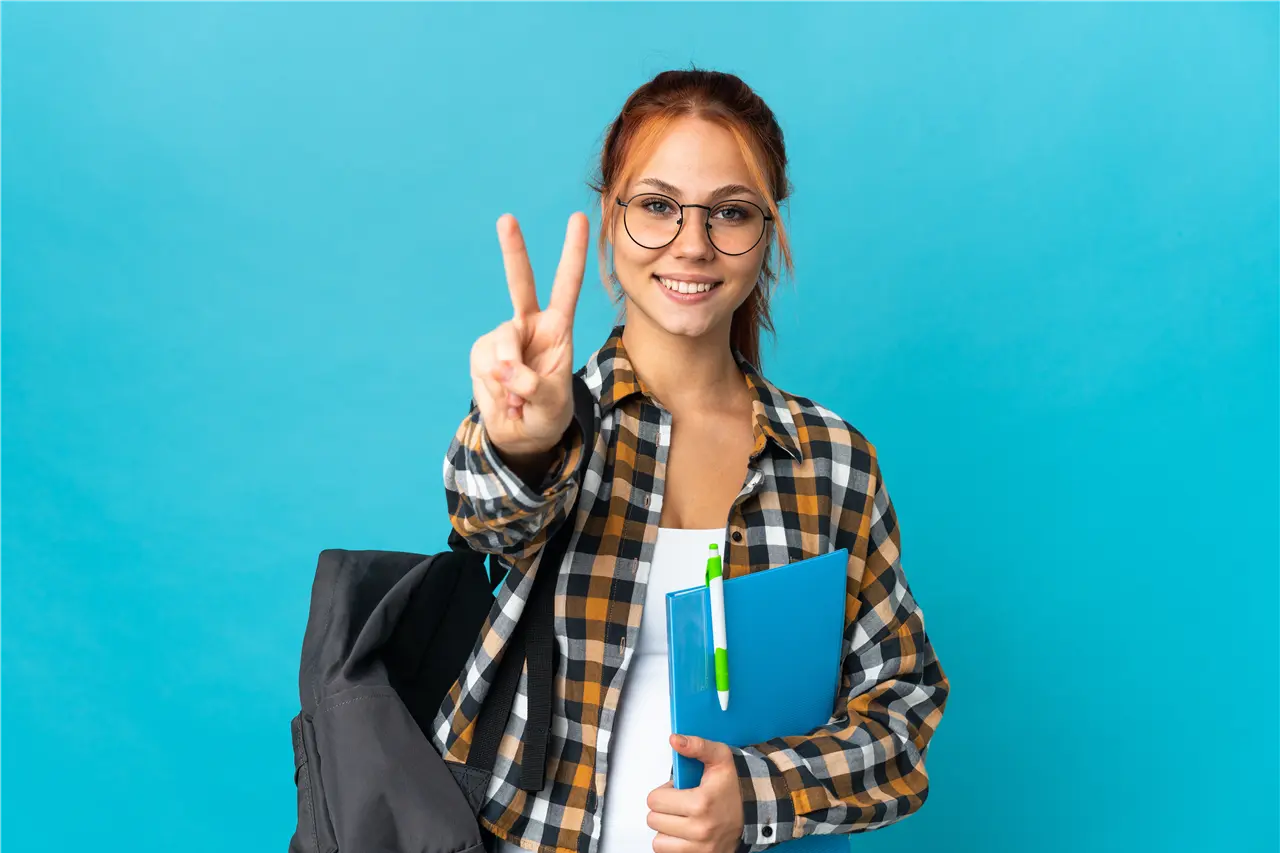 Eine Person zeigt das Peace-Zeichen und hält Ordner und Stift in der Hand. Gekleidet in ein kariertes Hemd und Rücken vor einem blauen Hintergrund trägt sie einen schwarzen Rucksack.