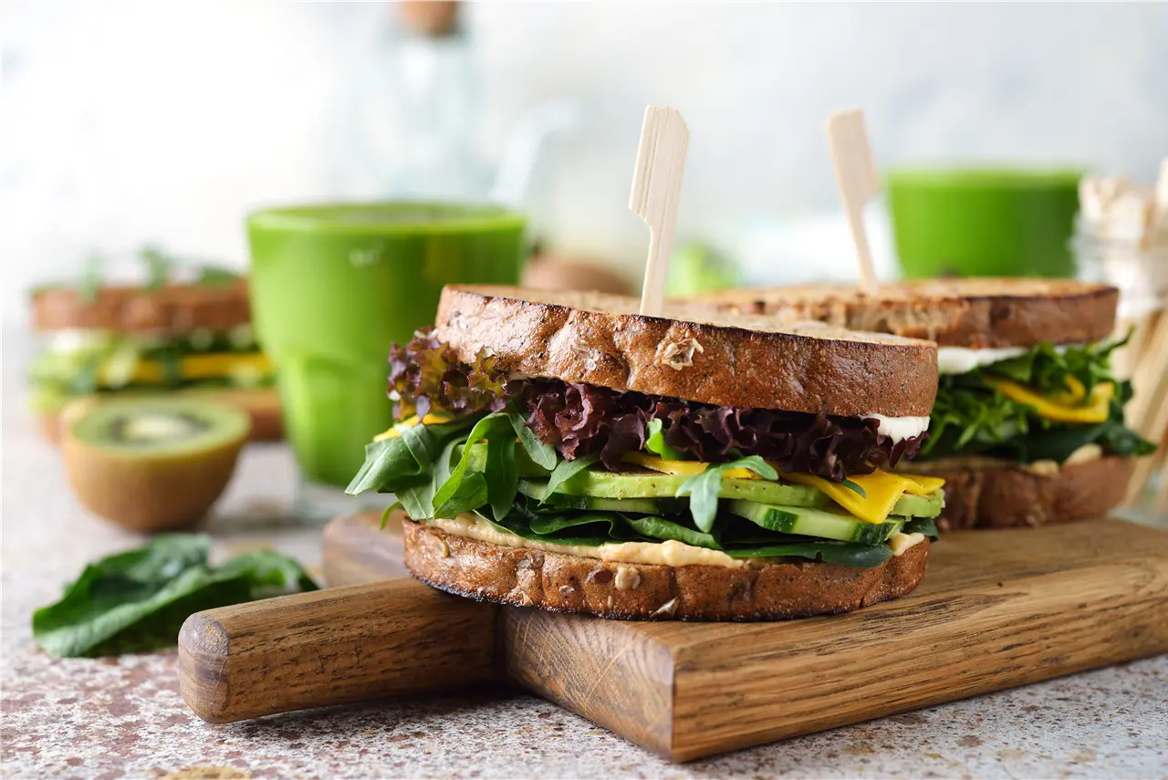 Zwei gesunde Sandwiches mit Vollkornbrot, gefüllt mit frischem Spinat, Gurken, Zucchini und Salat, werden auf einem Holzbrett serviert. Im Hintergrund sind ein Kiwi und grüne Smoothies zu sehen.