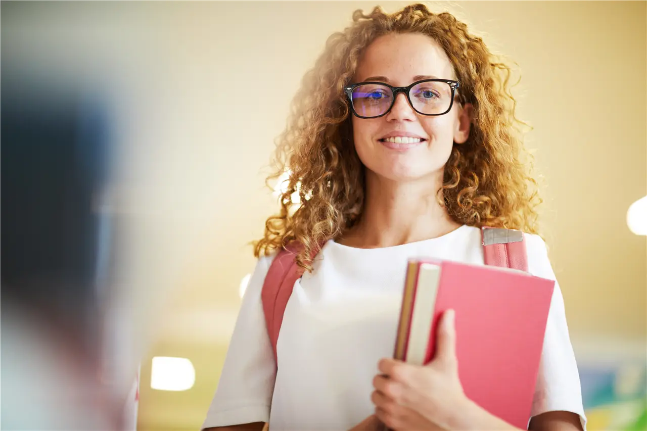 Eine Person steht mit einem Buch in den Händen und trägt einen Rucksack. Der Hintergrund ist verschwommen.