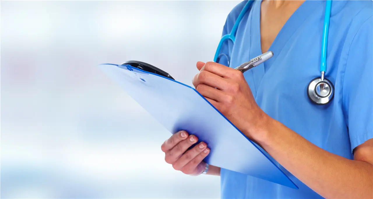Die Grafik zeigt eine Krankenschwester oder einen Arzt, die/der in blauer medizinischer Kleidung mit einem Stethoskop um den Hals auf einem Klemmbrett schreibt. Hintergrund ist leicht verschwommen.